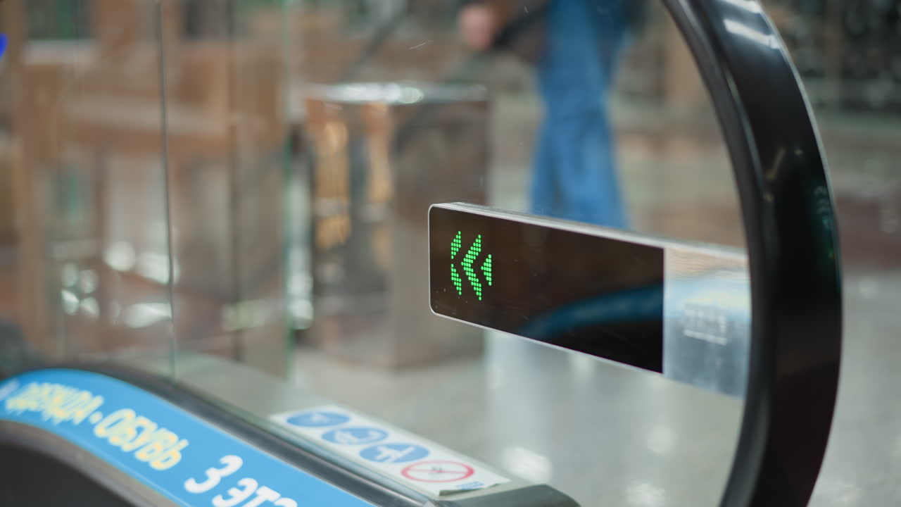 close up glass mounted green arrow led display indicating direction at entrance of moving walkway inside mall, pedestrians casting shadows on reflective surface under overhead lighting