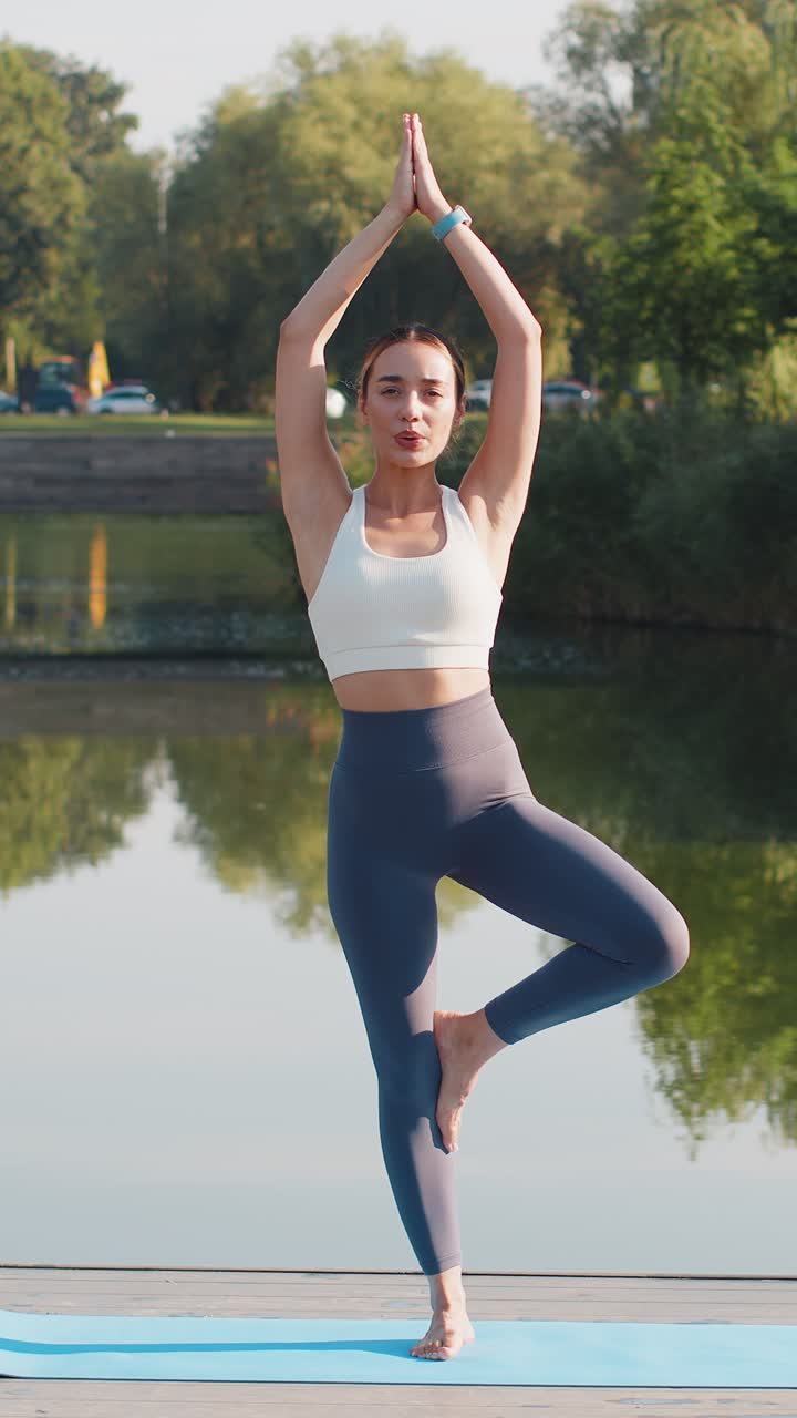 Jovem mulher atraente, instrutora esportiva, em roupa de ginástica, fazendo a postura da árvore de yoga perto de um lago no parque.