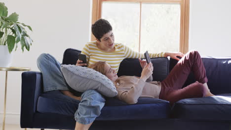 Feliz-Pareja-De-Lesbianas-Caucásicas-Tumbadas-En-El-Sofá,-Usando-Tabletas-Y-Teléfonos-Inteligentes-Y-Sonriendo-En-Una-Casa-Soleada