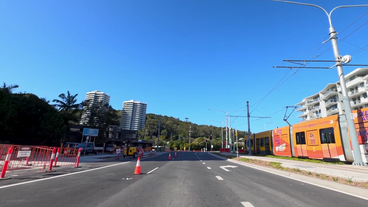 Perspectiva en primera persona desde un vehículo pasando por tranvías de tren ligero y edificios modernos bajo cielos azules despejados