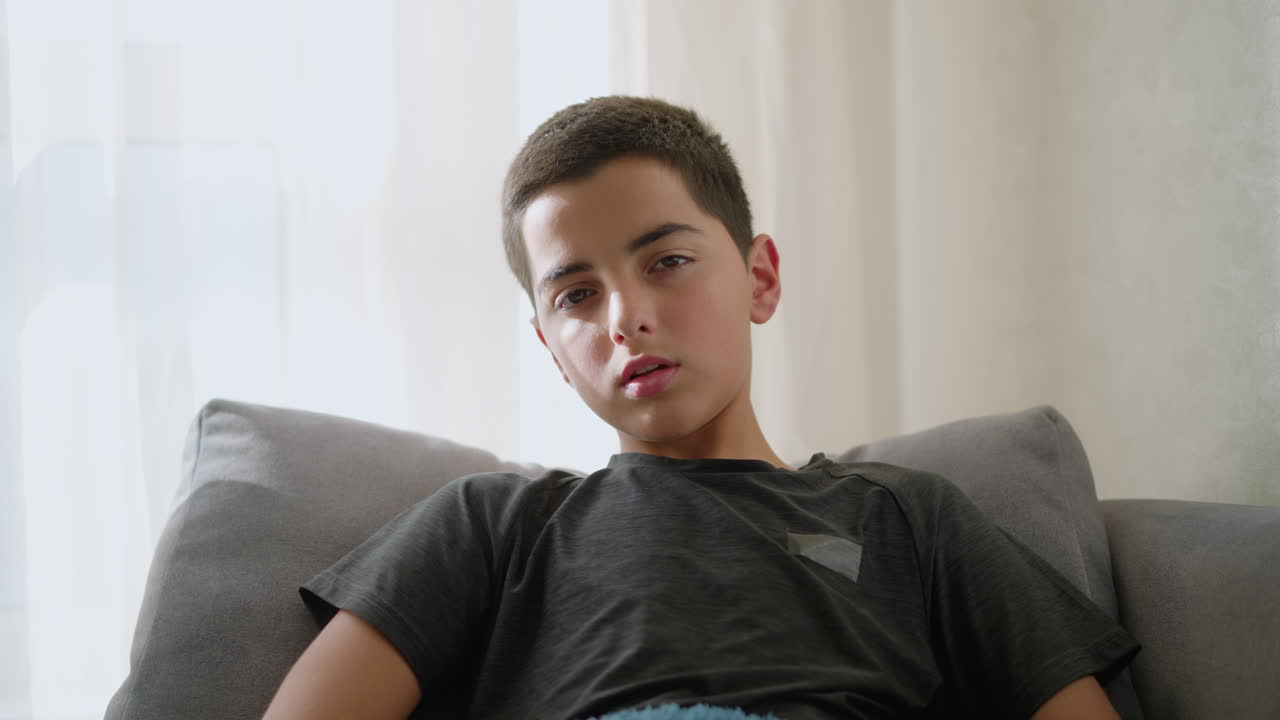 Portrait view of sick boy resting on the couch, looking weak and exhausted, wrapped in a blanket, with tissues scattered around, showing his tired, unwell state