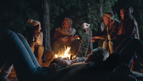 camping friends using smartphone browsing social media lying on ground by campfire at night in forest