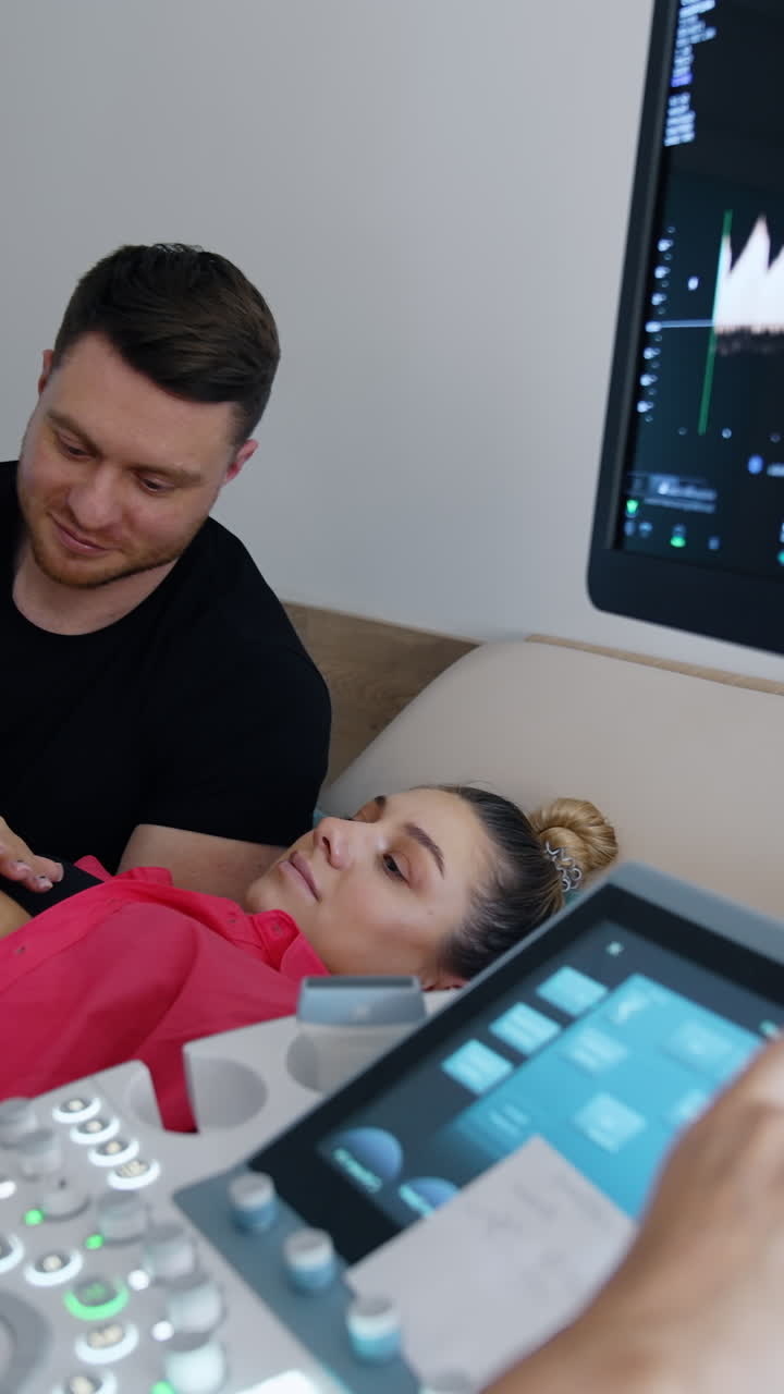 Ostetrica controlla la donna incinta con apparecchiature ecografiche. I futuri genitori guardano attentamente il monitor. Video verticale.