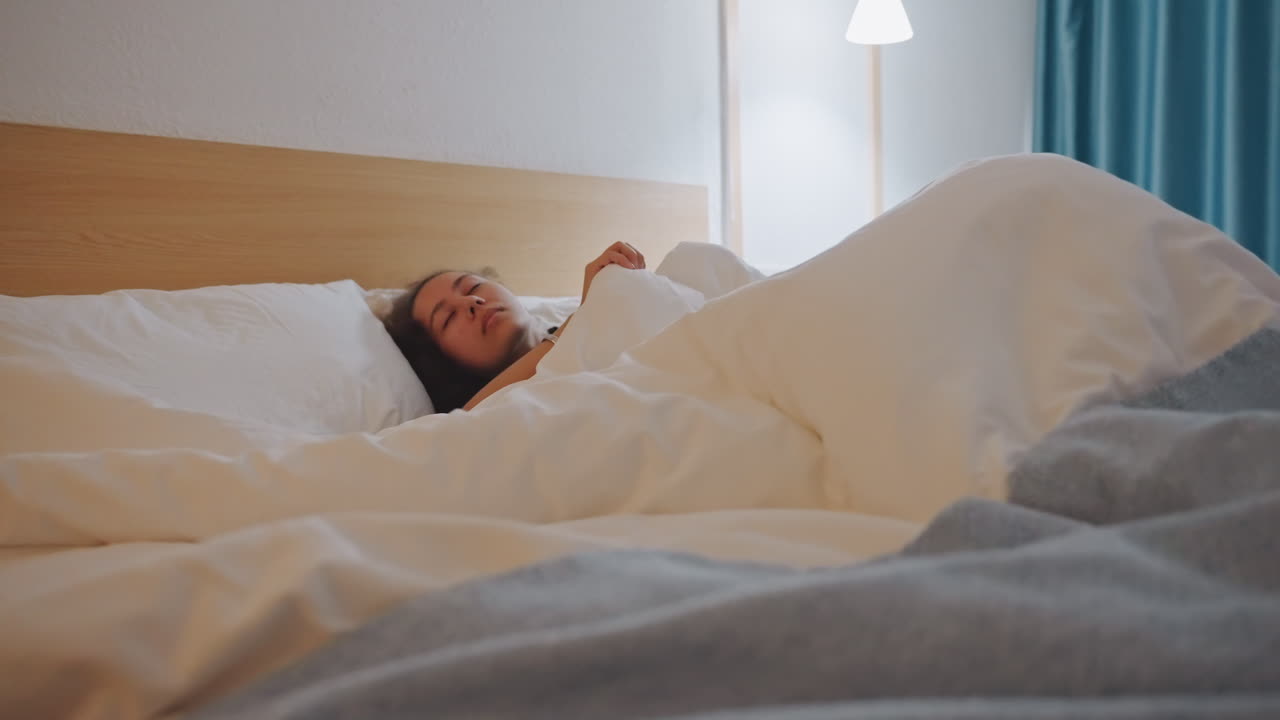 Kvinde vågner i hotelseng, strækker sig i hvid dyne, tjekker telefon, blødt lampeskær, blå gardiner, sengegavl af træ, afslappet morgenstemning, rejseatmosfære, omgivelser på boutiquehotel.