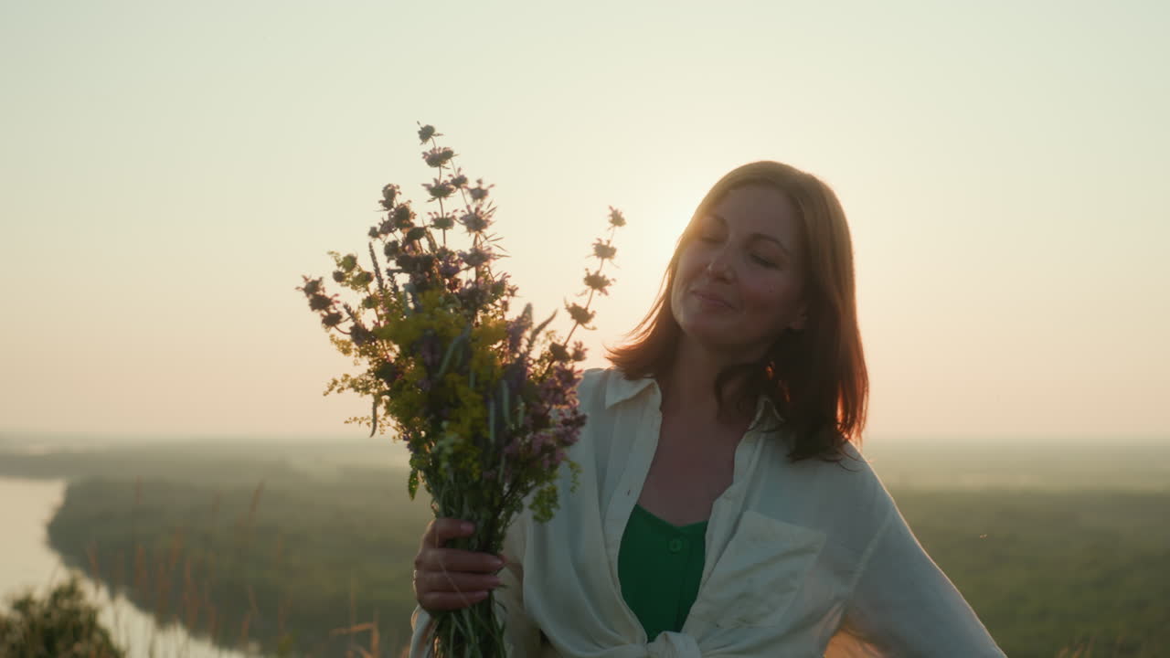 Sunset backlit woman in green dress spinning joyfully on grassy riverbank holding wildflower bouquet under sun flare above misty river valley glowing with golden light and gentle breeze