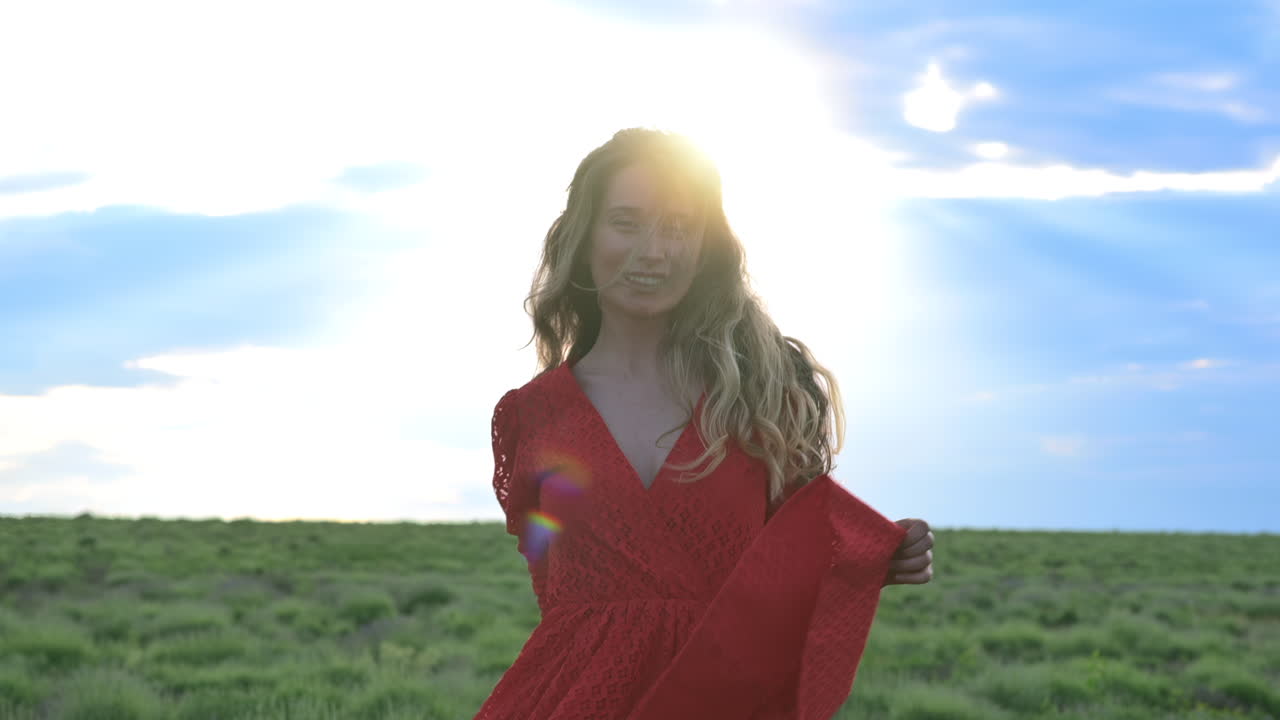 A woman in a red dress walks through a lush green field with the wind in her hair