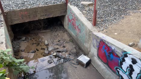 Graffiti-En-La-Pared-De-Cemento-De-Un-Canal-Con-Agua,-Piedra-Y-Arena.