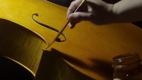intimate view of a luthier brushing varnish onto the golden wood of a cello’s top plate, focusing on the delicate f-hole curves, edges and purfling, in a traditional cremona setting