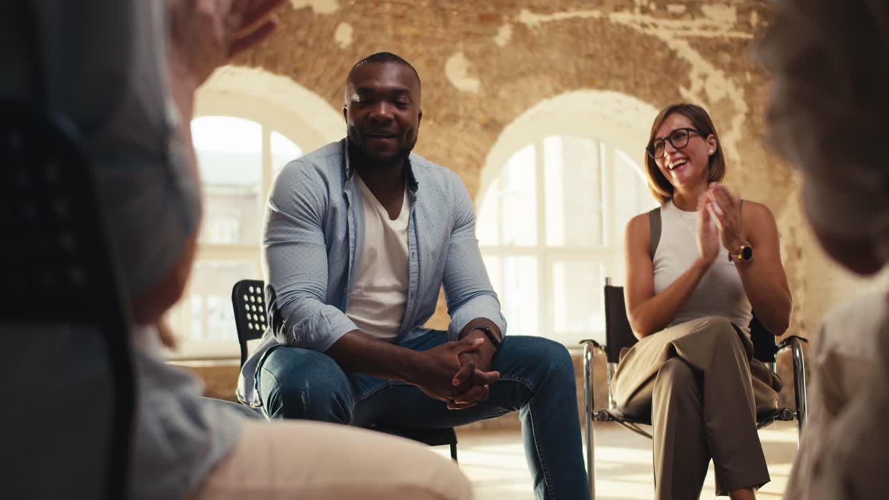 A Black man in a blue shirt expresses his thoughts in group therapy while other participants applaud him at a group therapy meeting in a yellow brick hall
