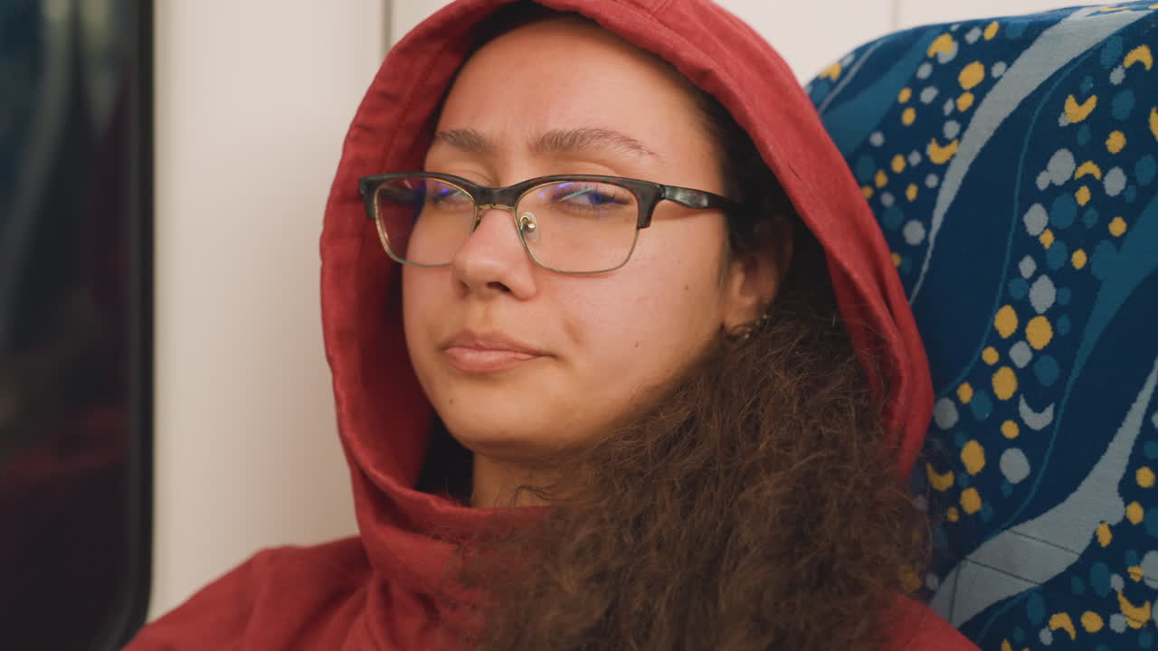 Donna con felpa con cappuccio e occhiali che riposa. Viaggiatrice rilassata con occhiali sul sedile del treno di sera. Donna serena con felpa rossa e occhiali che sonnecchia pacificamente in treno durante il tragitto serale.