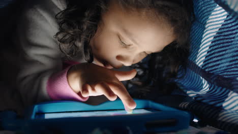 niña linda usando una tableta digital bajo una manta disfrutando de dibujar en la tecnología de pantalla táctil jugando juegos divirtiéndose antes de acostarse