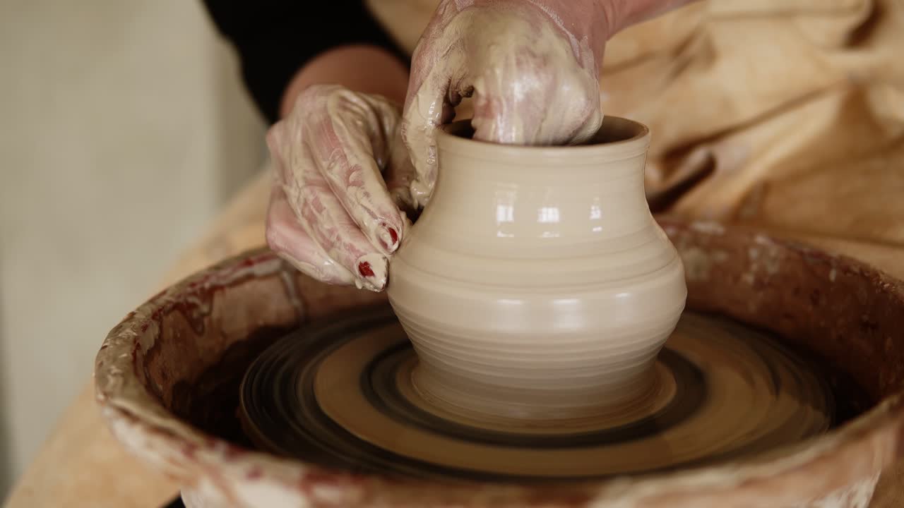 Female potter's hands with red manicure working with wet clay on a pottery wheel making a clay product in a workshop. Unrecognizable female person forming a vase, pulls it up