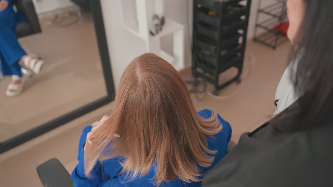 Peluquero en un salón peinando una melena a la altura de los hombros, primer plano desde atrás mostrando el espejo y la capa, peluquero seccionando las puntas y comprobando el largo, carro y herramientas de peluquería modernos al fondo, iluminación suave