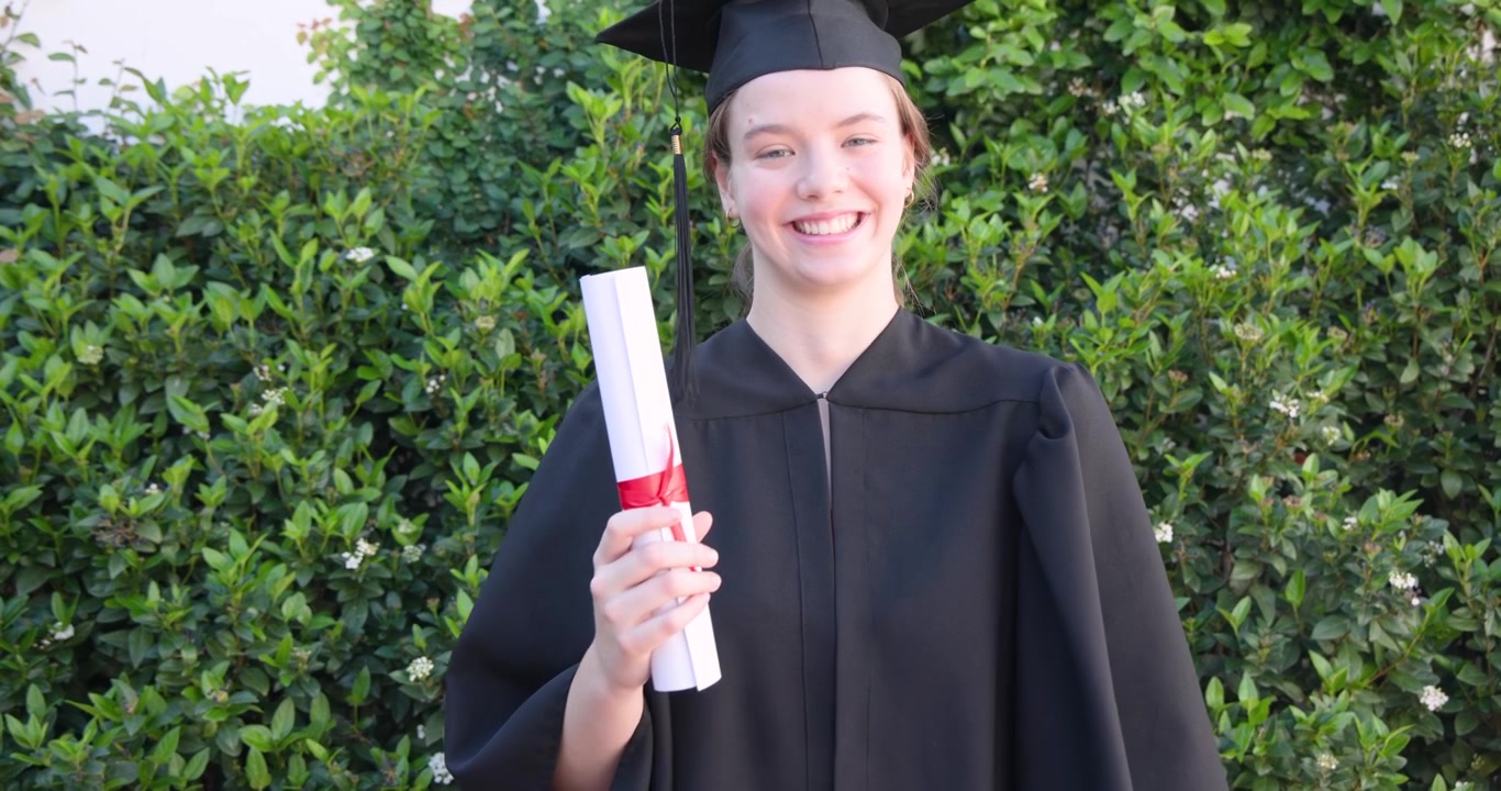 Female graduate after grad in gown cap taking phone selfies holding red ribbon diploma by hedge