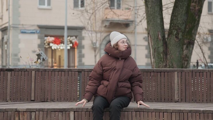 Caucasian woman commuter dazed on bench, regaining consciousness and scanning street for help winter parka and beanie, groggy movements and worried gaze near storefronts and muted traffic, intimate