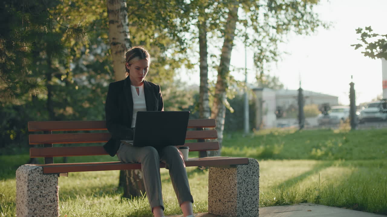 Contador remoto vestindo blazer focado em equilibrar a conta usando um laptop, sentado em um banco de madeira em um parque ao ar livre, com uma xícara de café ao lado e um plano de fundo urbano com edifícios