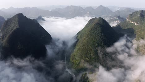 Asian-karst-mountains-rising-above-clouds-and-small-Chinese-town,-aerial