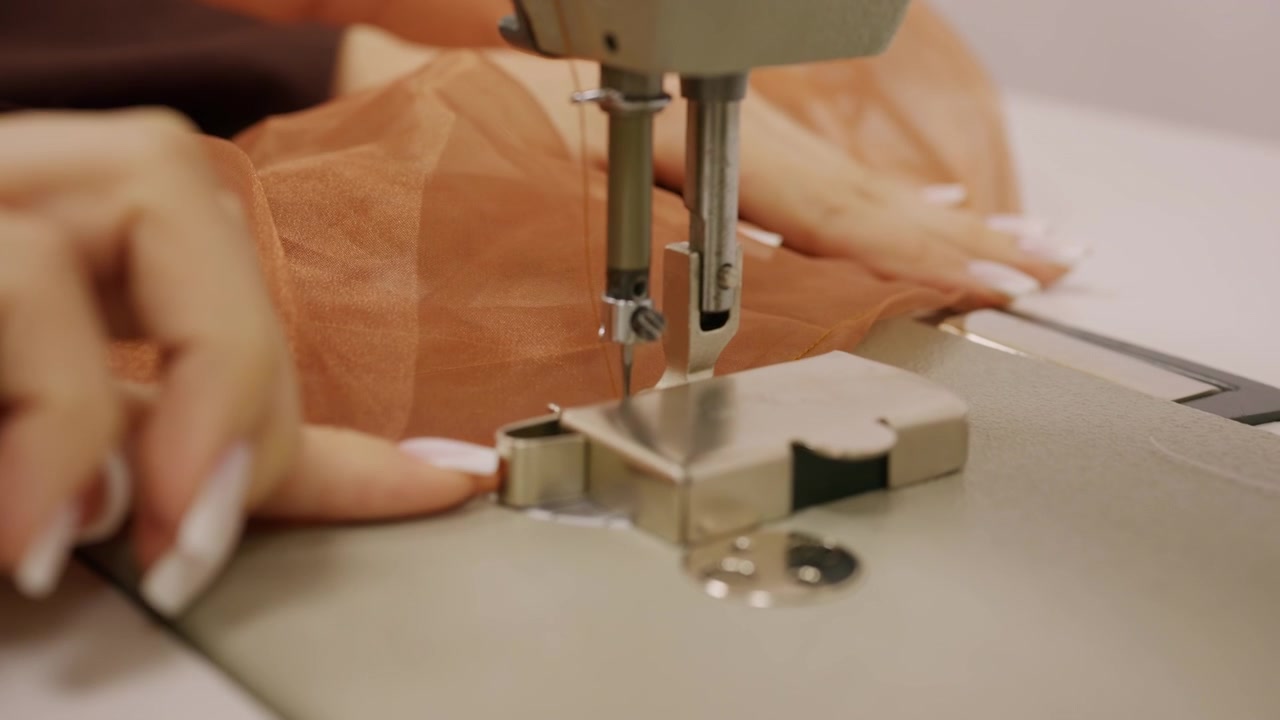 Plano detalhe de uma pessoa guiando tecido marrom transparente sob a agulha de uma máquina de costura industrial.