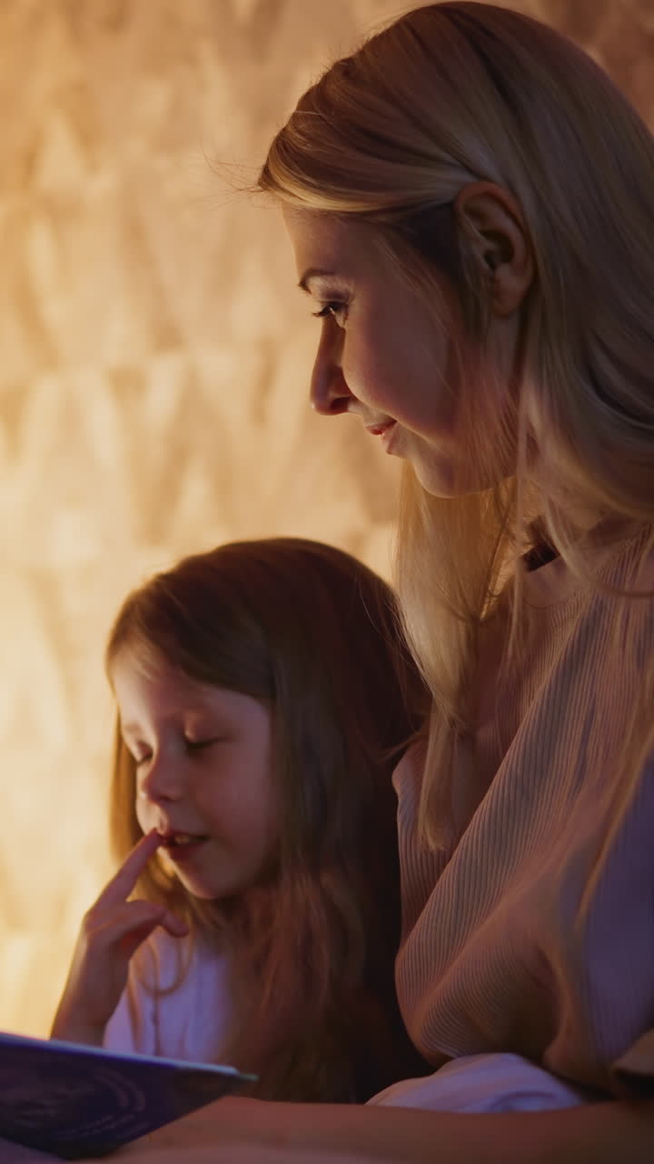 Little child girl touches lip with finger and reads book sitting near positive dimly lit mom on bed against play teepee in shaded bedroom closeup slow motion