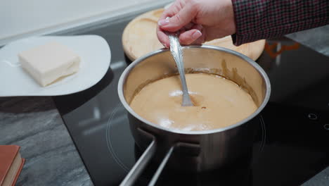 person holding pot on electric stove while stirring thick porridge as it bubbles due to heat, partial view of white plate with butter and wooden board on dark countertop in modern kitchen setting