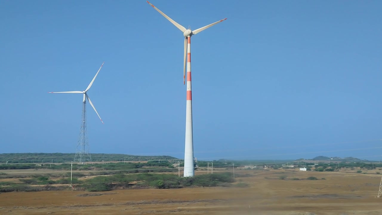 Vista da un veicolo in movimento di grandi turbine eoliche in un paesaggio arido sotto un cielo sereno, ideale per progetti di energia rinnovabile, sostenibilità, clima e infrastrutture industriali.