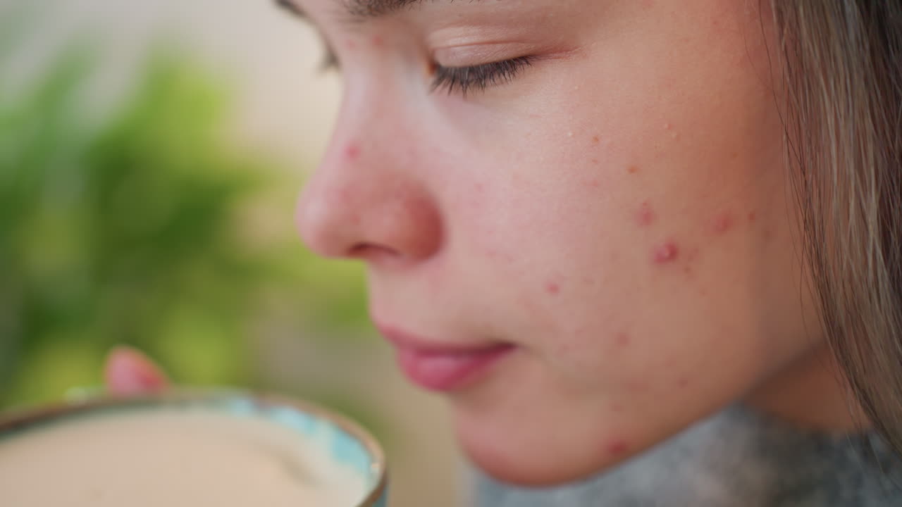 Close view of young lady with facial spots gently perceiving scent from breakfast beverage held with both hands subtle smile on lips satisfied expression blurred flower decor in background