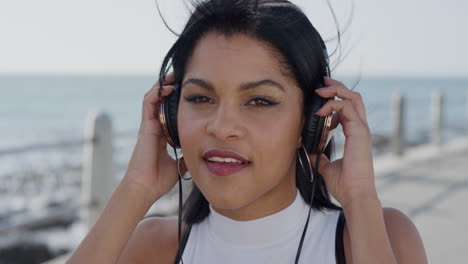 portrait beautiful young hispanic woman wearing headphones dancing enjoying listening to music wind blowing hair relaxing on warm seaside ocean background