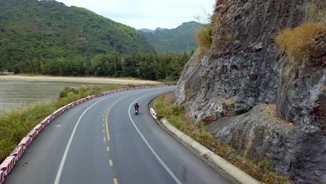 woman on a moped riding on an island road near the coastline, aerial follow behind shot