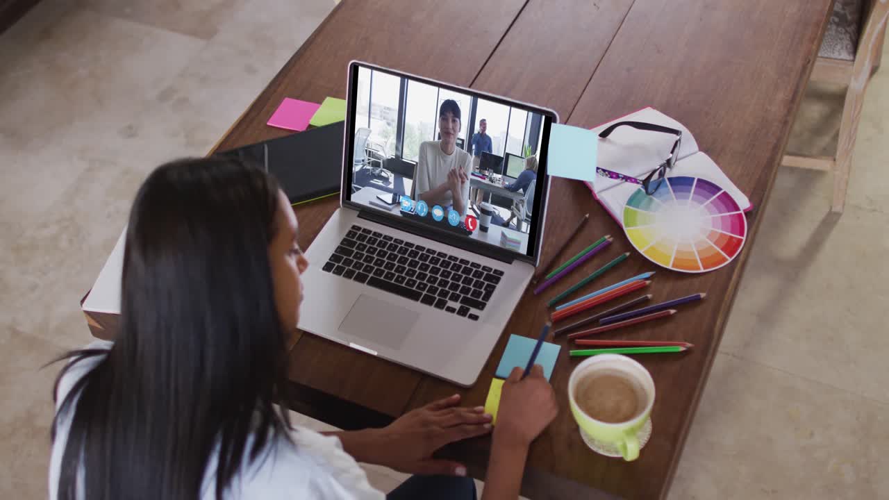 Mujer caucásica usando una laptop en videollamada con una colega y tomando notas