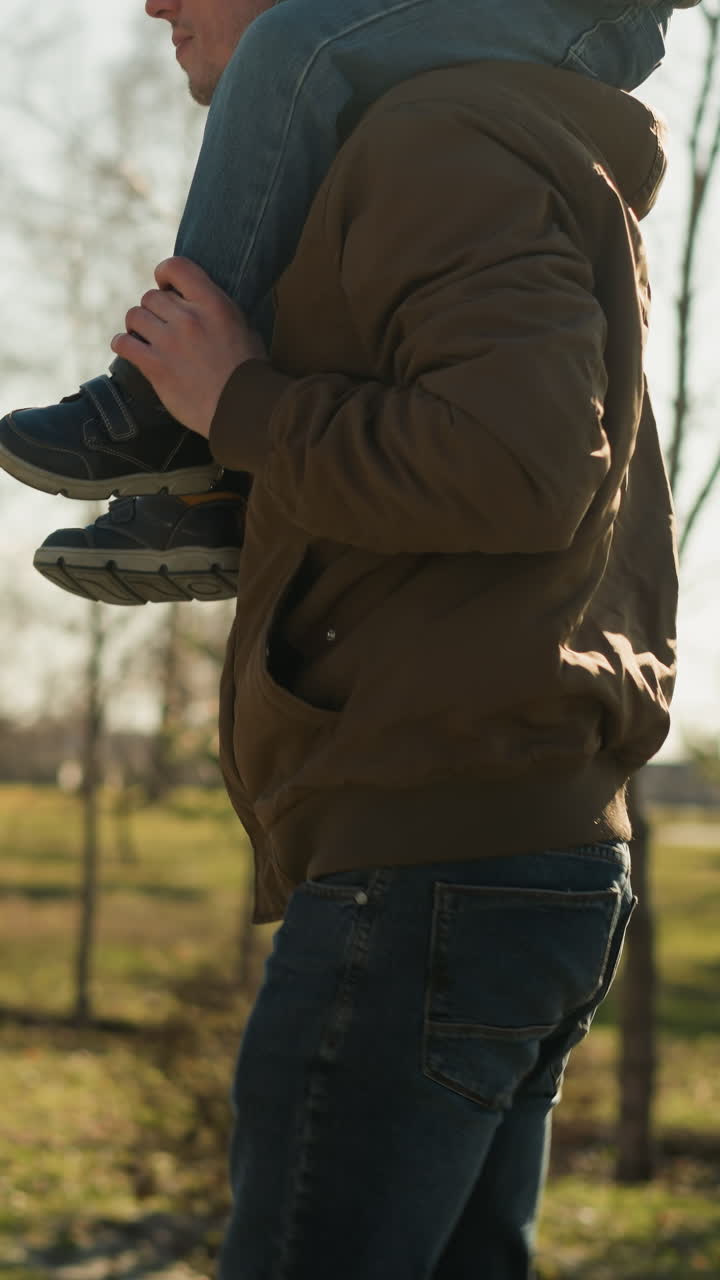 A close-up of a father walking with his son on his shoulders, focused on where they are headed, as they walk on a paved path near an open field, the father wears jeans and black sneakers