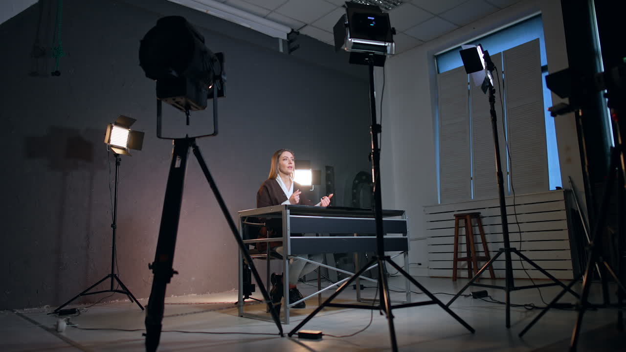 Soffits directed on the woman sitting at desk and talking. Low angle view at the confident lady recording blog in the studio