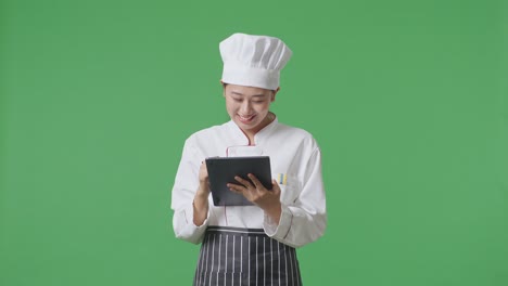 Asian Woman Chef Taking Note On A Tablet While Standing In The Green Screen Background Studio