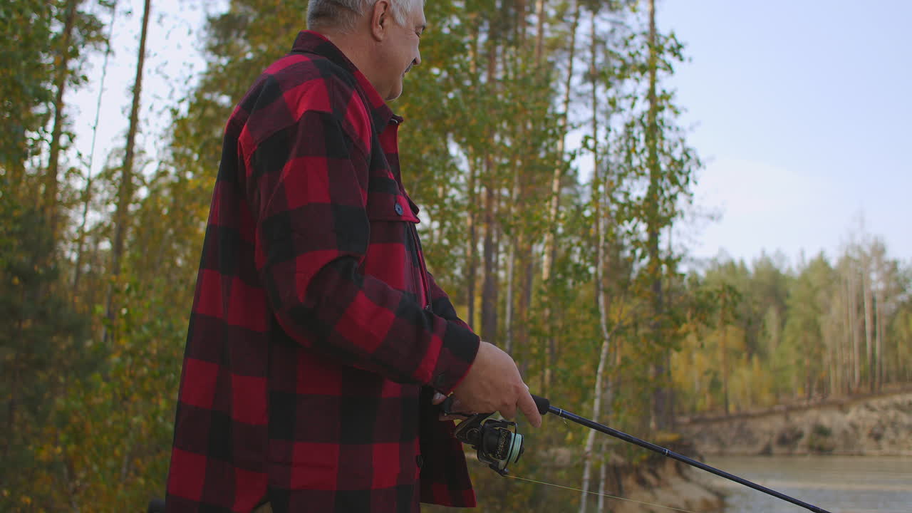 adult fisherman is rotating reel of rod during spin fishing in freshwater standing on shore of river in forest at autumn day