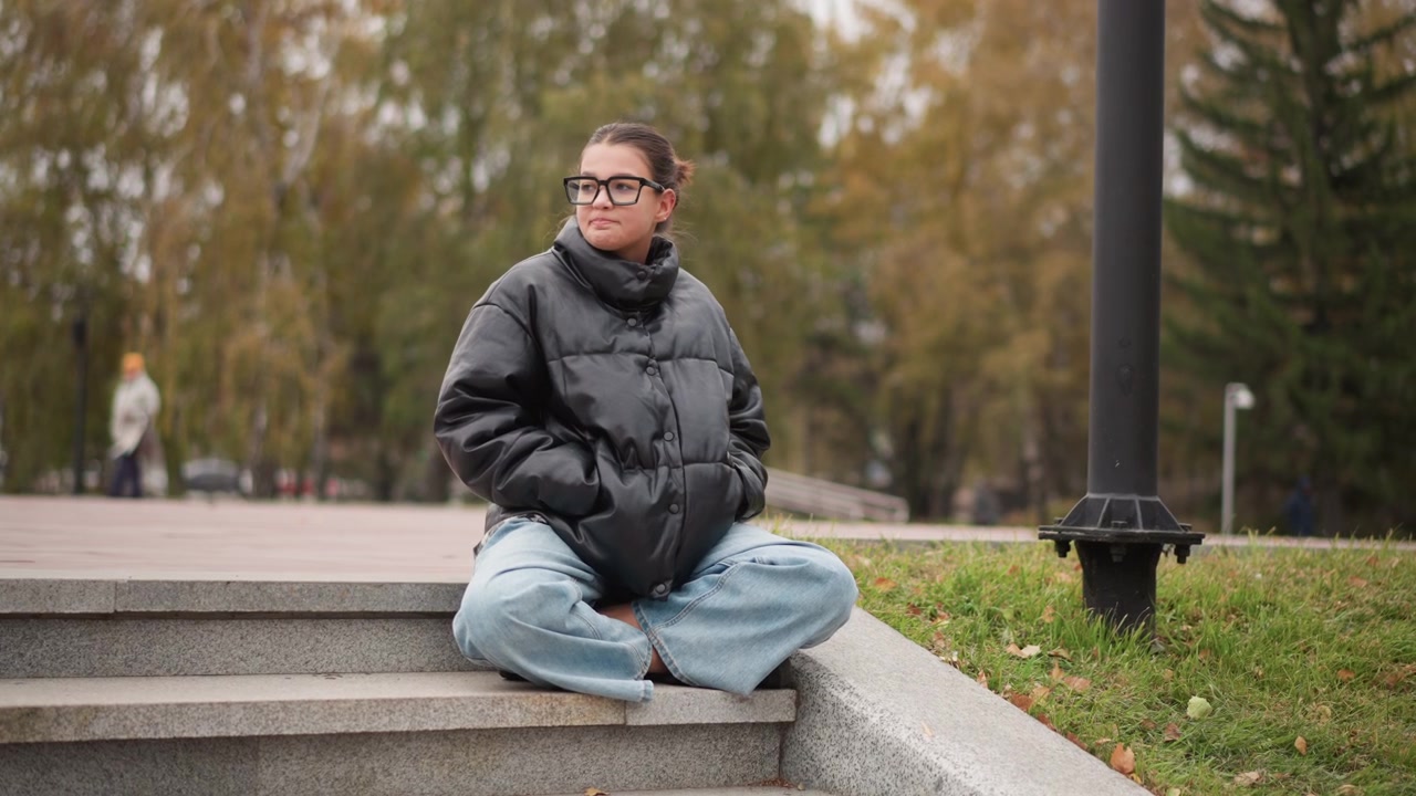 Young woman sits cross legged on stone steps in outdoor park setting wearing black puffer jacket and light blue jeans. scene features soft natural lighting with autumnal trees visible