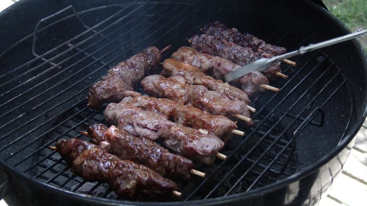 Membalik kebab daging sapi dan babi marinasi di atas panggangan menggunakan penjepit di atas api terbuka.