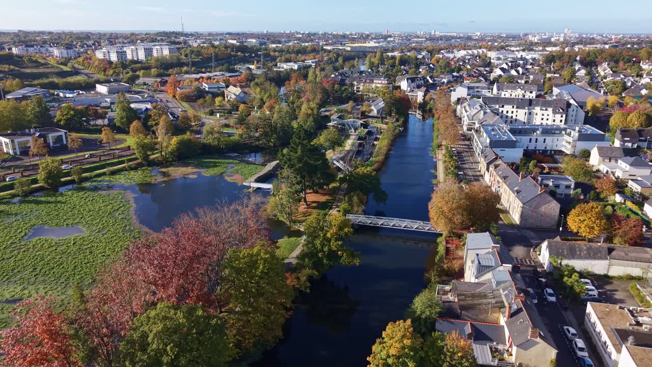Aerial forward-moving drone shot over La Vilaine River in Cesson-Sévigné, showing autumn-colored trees, a small bridge, and nearby houses