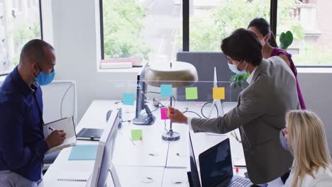 Diverse-business-colleagues-wearing-face-masks-using-computers-talking-in-office