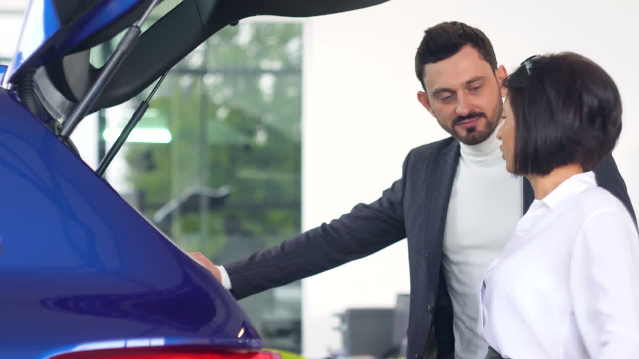 Professional car female salesman is telling interested buyer stylish man about luxurious car with trunk open in motor show