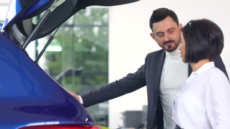 professional car female salesman is telling interested buyer stylish man about luxurious car with trunk open in motor show