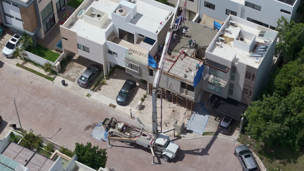 Panning tilt down drone shot of a concrete pump pouring cement to make the tile of a house with several workers spreading the material