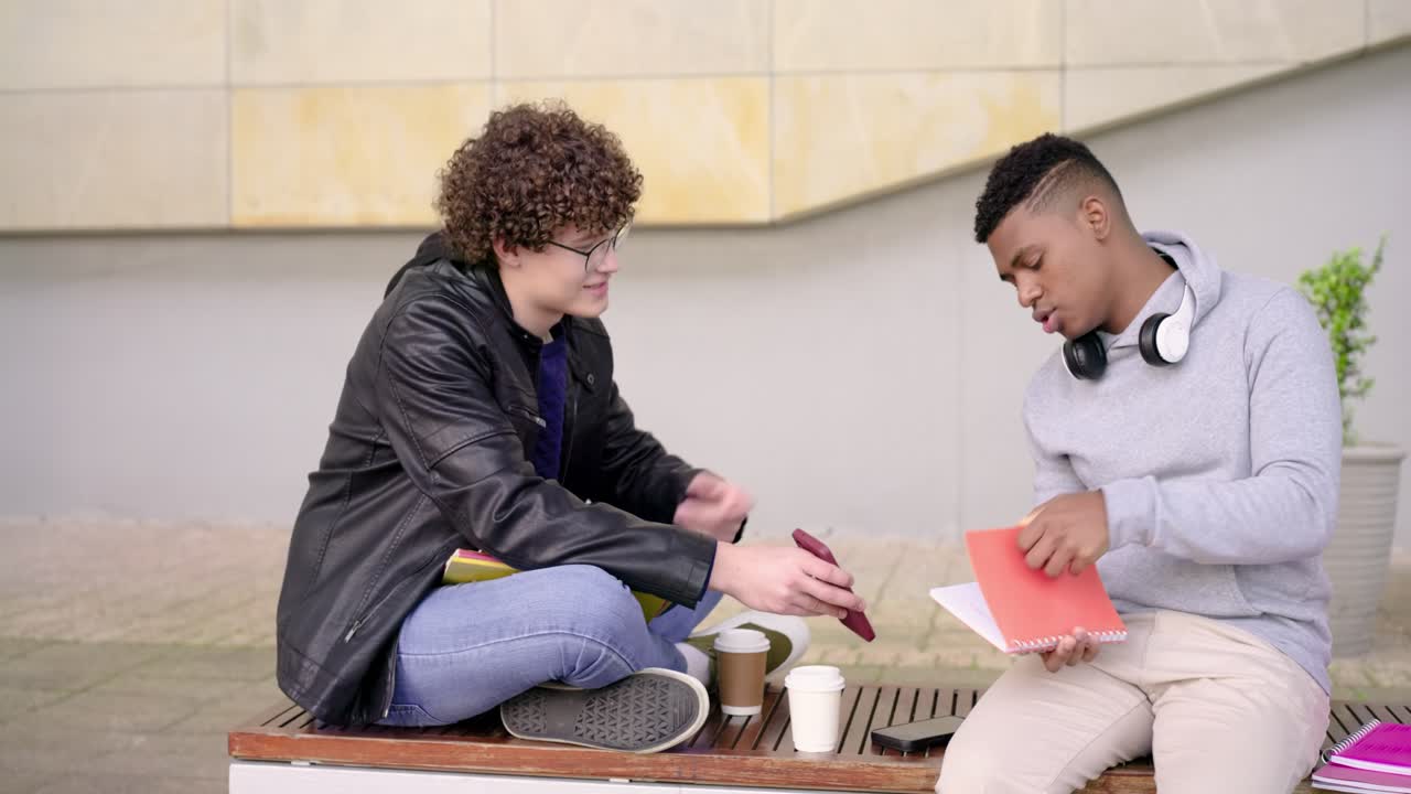 Estudantes masculinos diversos num banco, estudante de cabelo encaracolado a levantar o olhar do telemóvel, mostrando uma pasta enquanto estuda.