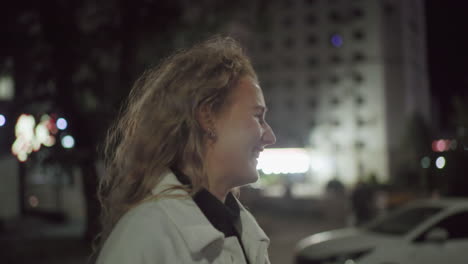 shy smiling woman in white coat looking away from camera under bright night lights in urban cityscape with blurred building and colorful bokeh lights in soft background