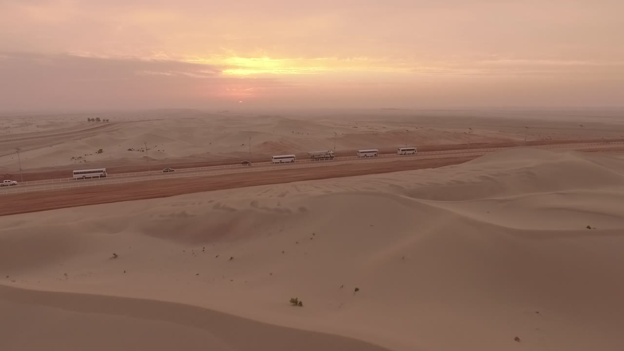 Aerial Shot of a Sunset in the Abu Dhabi Desert, the Camera Glides Over the Dunes Towards the Sun, Approaching a Sandy Road Where Vehicles and Buses Travel in Line.