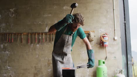 Caucasian-male-blacksmith-wearing-safety-glasses,-hammering-hot-metal-tool-on-anvil-in-workshop