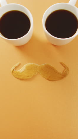 video vertical de dos tazas de café y bigote falso rubio en fondo naranja con espacio de copia
