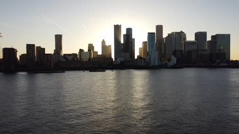 Aerial-drone-flight-over-River-Thames-showing-silhouette-of-skyscraper-buildings-in-Canary-Wharf-District-of-London-during-sunrise