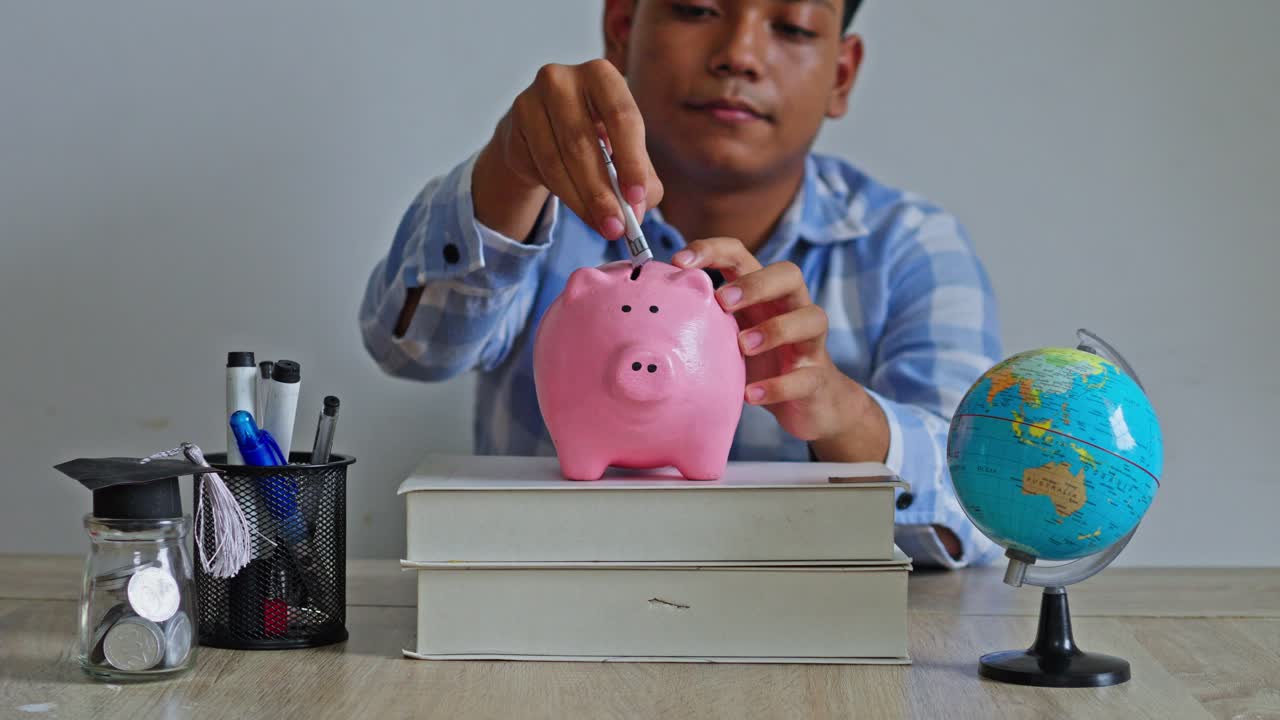 Student inserting cash into money box with globe and graduation-themed jar on desk