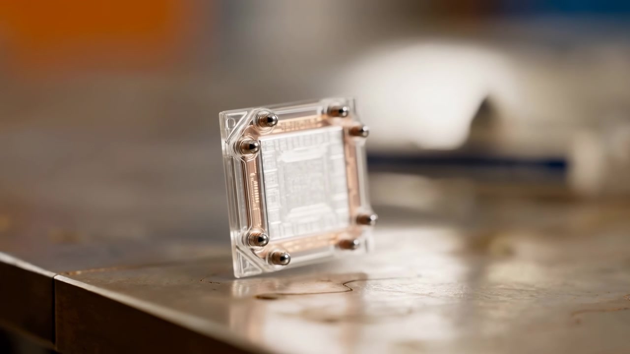 Vista ravvicinata di un componente elettronico rettangolare trasparente con canali interni in rame e sei piedini metallici arrotondati in posizione verticale su un tavolo in legno usurato o laminato, illuminato delicatamente per un aspetto pulito.