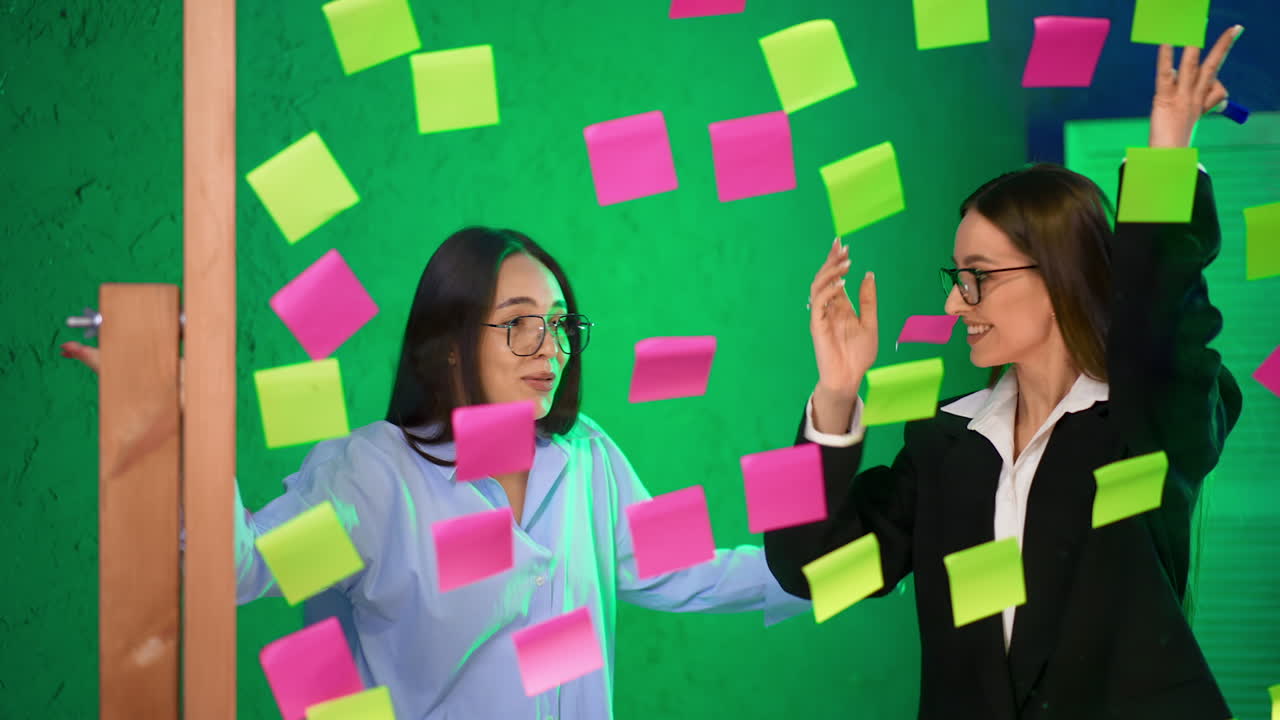 Happy beautiful brunette Caucasian ladies stand behind the glass wall. Women take the stickers of the board and throw them back.