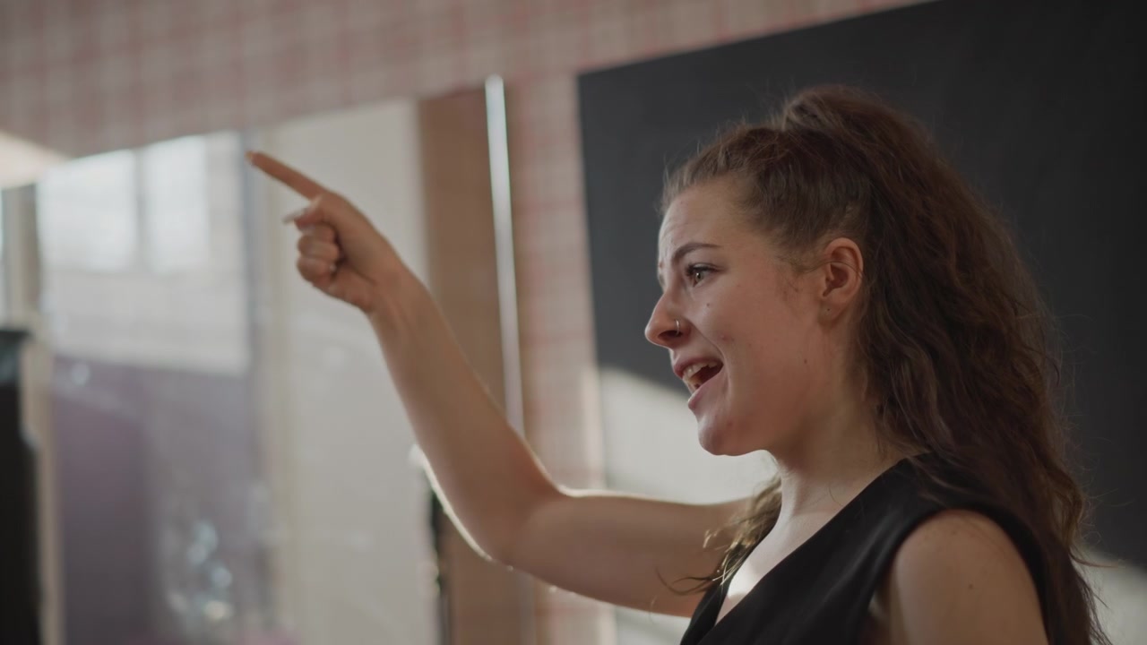Ung vit kvinna leder sångövning, energisk masterclass-känsla när sångaren pekar och vägleder studenter, auktoritära gester och timingövningar i en studio-klassrum, självsäkert ledarskap